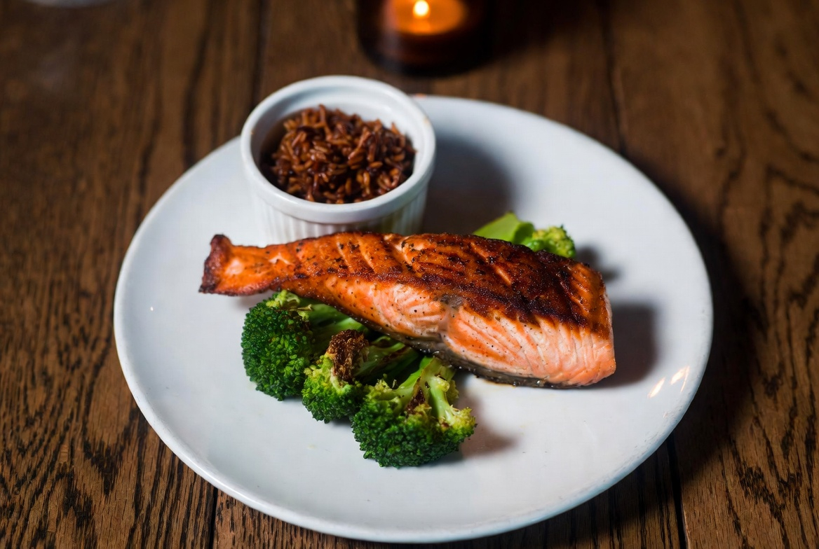 Split plate composition showing grilled salmon fillet, steamed broccoli, and brown rice, white plate, restaurant plating style, warm lighting