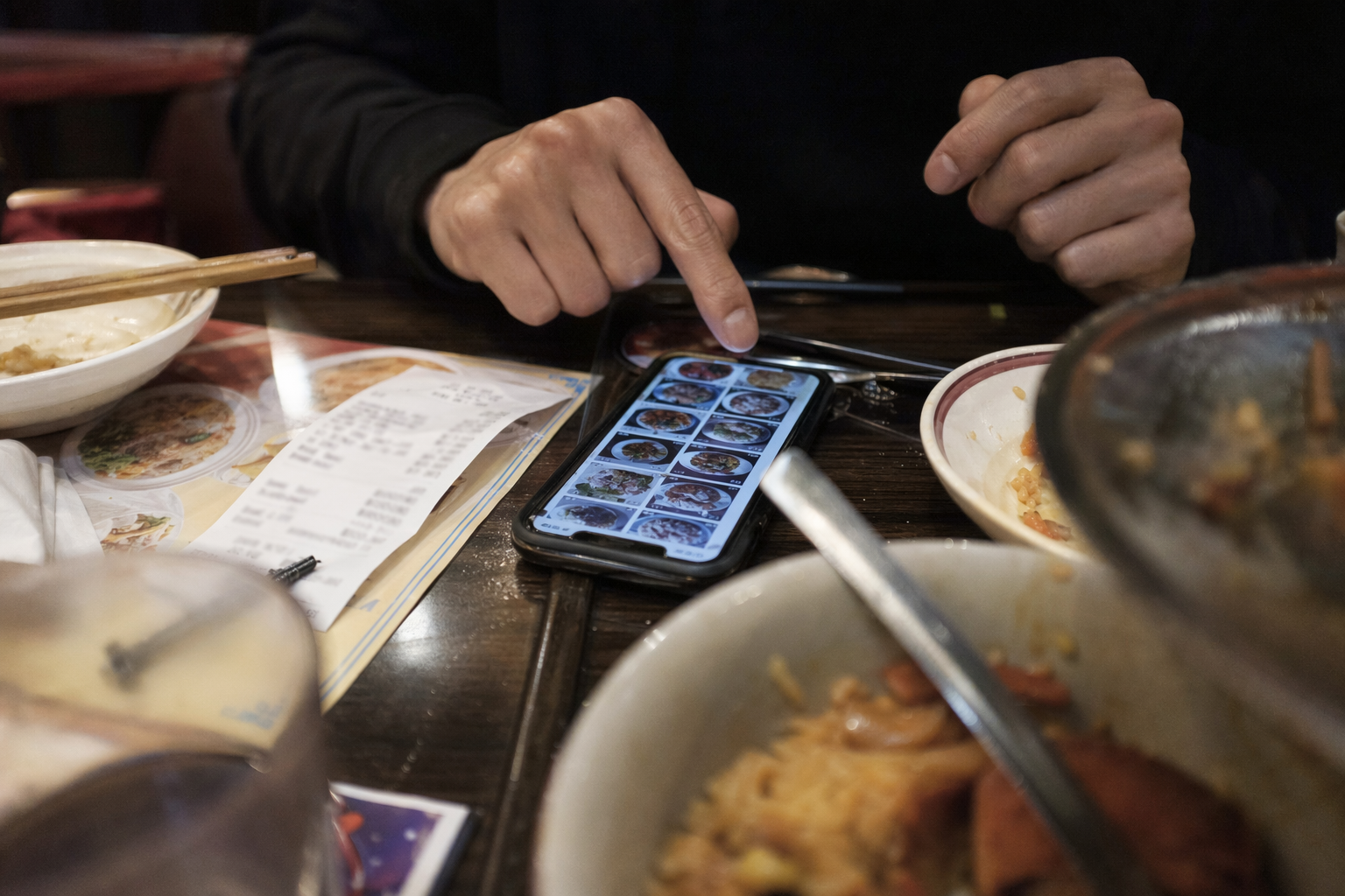 Tight close-up of a phone on a busy restaurant table beside a receipt, menu, and partially visible dishes, realistic editorial lifestyle photograph